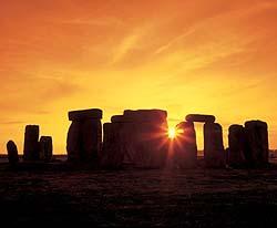 Shows a photo of the standing stones at Stonehenge, in silhouette against an orange sky, with the sun peeking through them.
