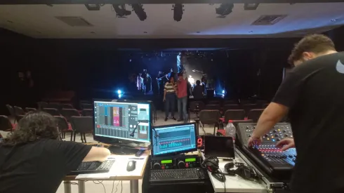 Monitores y mesas de sonido en un primer plano; en el segundo el profesor con una alumna y, al fondo, el escenario  donde se están montando monitores, micrófonos y espacios para los músicos. 