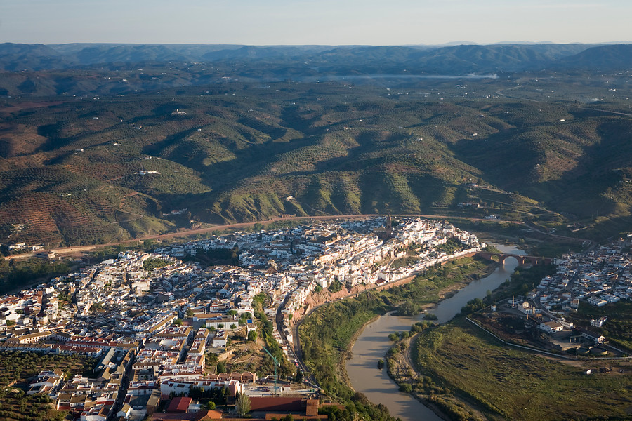 Ampliar imagen: fotografía aérea de un pueblo de casa blancas a la izquierda de la imagen, río Guadalquivir pasando por la derecha del pueblo y montañas verdes  en el resto de la imagen