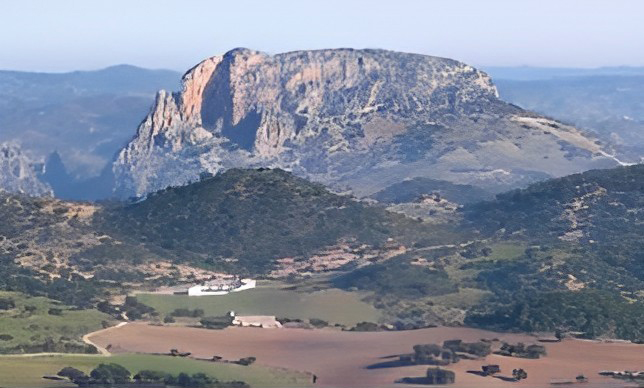 Panorámica del peñón rocoso de Zaframagón. Primer plano de suaves laderas con arboleda, cultivos, grupos de árboles dispersos y edificios