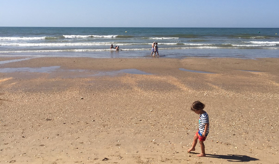 Un niño juega en la arena de una playa andaluza con paseantes y bañistas al fondo.