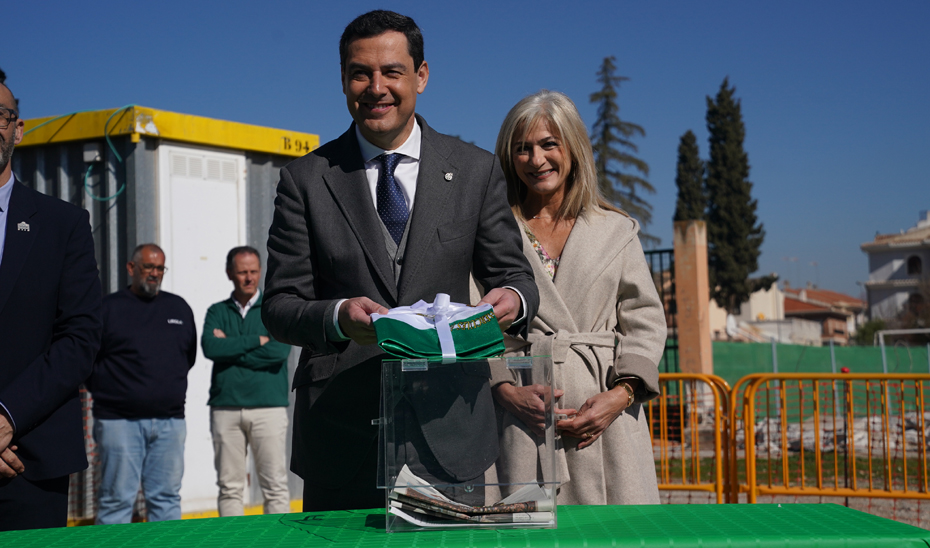 Juanma Moreno introduce la bandera de Andalucía en la caja con documentos y objetos representativos utilizada en el acto de colocación de la primera piedra del futuro instituto.